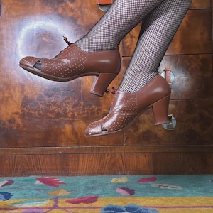 Puede incluir: Zapatos peep-toe de cuero marrón con pequeños lunares blancos. Los zapatos tienen un tacón grueso y detalles de cordones. La persona lleva medias de rejilla negras. El fondo es un armario de madera y una alfombra colorida.