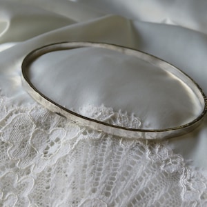 Puede incluir: Un brazalete de plata con acabado martillado, descansando sobre un tejido de encaje blanco.