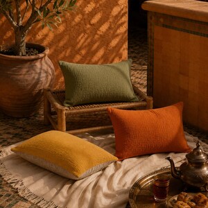 May include: Three rectangular throw pillows in solid olive green, burnt orange, and mustard yellow colors. Each pillow features a textured, crinkled fabric surface. The pillows are arranged on a small wooden stool and a cream-colored fringed throw.