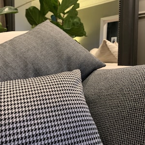 Puede incluir: Tres almohadas decorativas con diferentes patrones. La almohada superior es de color gris oscuro con un patrón de espiga. La almohada del medio es de color blanco y negro con un patrón de pata de gallo. La almohada inferior es de color gris oscuro con un pequeño patrón texturizado.