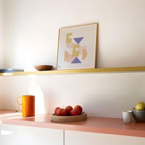 Könnte beinhalten: Moderne Innenraum-Szene mit einem gerahmten Kunstdruck mit abstrakten geometrischen Formen in Pastellfarben. Ein orangefarbener Becher, eine Holzschale mit roten Äpfeln und eine kleine weiße Tasse stehen auf einem rosa Regal.
