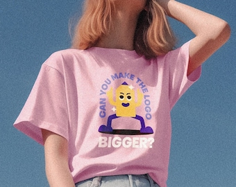 Könnte beinhalten: Ein pinkfarbenes T-Shirt mit einer Comicfigur und dem Text "Can you make the logo bigger?" darauf gedruckt. Die Person, die das Shirt trägt, sitzt auf einer Betonfläche.