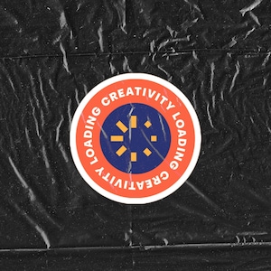 May include: A red and white circular sticker with a blue center. The sticker has the words "LOADING CREATIVITY" written around the outside. The center of the sticker has a stylized sunburst design.