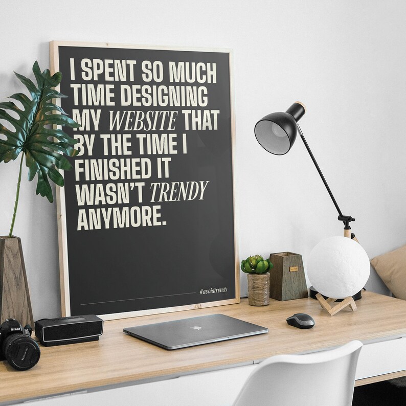 May include: A black and white poster with a quote about website design. The quote reads "I spent so much time designing my website that by the time I finished it wasn't trendy anymore." The poster is framed in wood and is hanging on a white wall.
