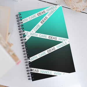 May include: A black and teal notebook with a spiral binding. The cover has white tape with the text "With Care Ideas Handle" repeated across it.