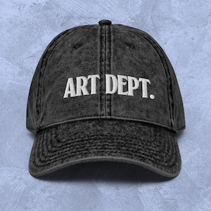 May include: Black vintage style baseball cap with white embroidered text that reads "ART DEPT."