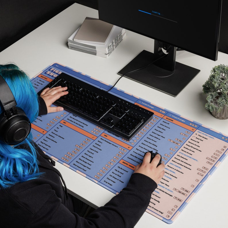 May include: A large rectangular mousepad with a blue and orange design featuring text and a keyboard. The mousepad is on a white desk with a black keyboard and mouse. A computer monitor and small plant are also visible.