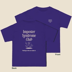 K&ouml;nnte beinhalten: Lilafarbenes T-Shirt mit dem Text "Imposter Syndrome Club" und einer Comicfigur auf der R&uuml;ckseite. Die Vorderseite des Shirts tr&auml;gt den Text "Imposter Syndrome Club" in kleinerer Schrift.