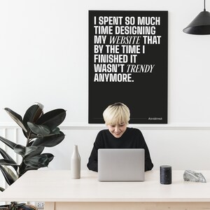 May include: A black and white poster with the text "I spent so much time designing my website that by the time I finished it wasn't trendy anymore." The poster is hanging on a white wall. A person is sitting at a table with a laptop in front of them.
