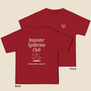 K&ouml;nnte beinhalten: Rotes T-Shirt mit einem wei&szlig;en Aufdruck auf der R&uuml;ckseite, der "Imposter Syndrome Club" mit einer Comicfigur und dem Text "Faking it till we make it." lautet. Die Vorderseite des Shirts hat einen kleinen wei&szlig;en Aufdruck mit der Aufschrift "Imposter Syndrome Club".