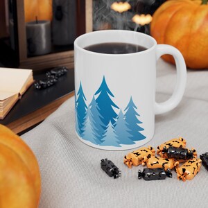 May include: White ceramic mug with a blue and white winter forest design. The mug is filled with coffee and has steam rising from it. The mug is sitting on a gray surface with orange and black candy.