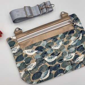 May include: A small crossbody bag with a beige fabric exterior featuring a pattern of blue and white birds on a background of gray and beige pebbles. The bag has a gold zippered pocket and a silver adjustable strap.