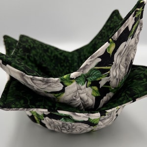 Puede incluir: Un juego de dos calientaplatos de tela negra y verde con un patrón floral blanco. Los calientaplatos están doblados en forma de cuenco.