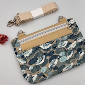 Op de afbeelding: Een klein beige en blauw crossbody tasje met een ritssluiting en een patroon van vogels en kiezelsteentjes. De tas heeft een lange verstelbare band.