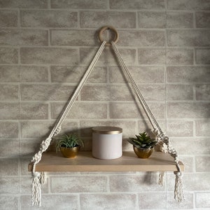 May include: A wooden shelf with a rope and knot design hangs from a ceiling hook. The shelf is decorated with two small potted plants and a white candle in a gold container.