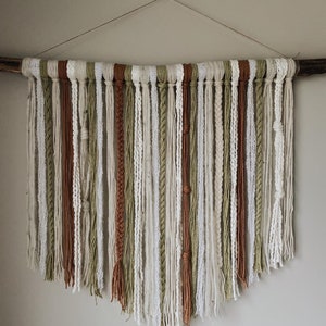 May include: A wall hanging made of various strands of yarn in shades of white, cream, brown, and green. The yarn is tied to a branch and hangs in a cascading pattern.