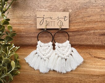 May include: White macrame earrings with gold hoops and tassels. The earrings are hanging from a brown card with the text "just britt co."
