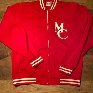 Puede incluir: Chaqueta roja con cremallera con rayas blancas en el cuello, los puños y el dobladillo. La chaqueta tiene un logotipo "MC" blanco en la parte delantera.