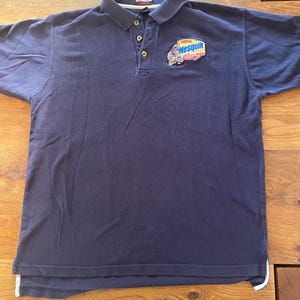 Puede incluir: Una camiseta polo azul marino con un logotipo de Nesquik que presenta un personaje de dibujos animados. El logotipo está bordado en el pecho izquierdo de la camiseta.