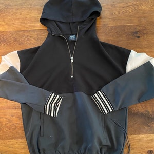 Könnte beinhalten: Ein schwarzer, weißer und grauer Kapuzenpullover mit Reißverschluss und Kordelzug am Saum. Der Pullover hat einen weißen Streifen am Ärmel.