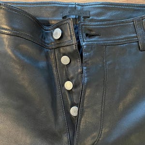May include: Close-up of black leather trousers with silver button fastenings. The trousers have a classic five-pocket design and a straight leg cut. The buttons are embossed with a circular design. The trousers are made of a smooth, shiny leather material.