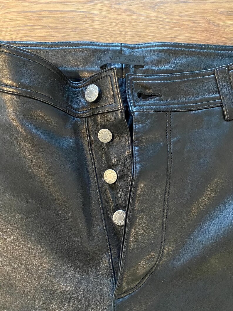 May include: Close-up of black leather trousers with silver button fastenings. The trousers have a classic five-pocket design and a straight leg cut. The buttons are embossed with a circular design. The trousers are made of a smooth, shiny leather material.