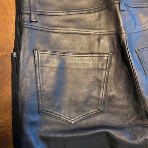 May include: Close-up of black leather trousers. The trousers have a classic five-pocket design with a back pocket visible. The leather appears smooth and has visible stitching details. The trousers have a belt loop and a button closure.