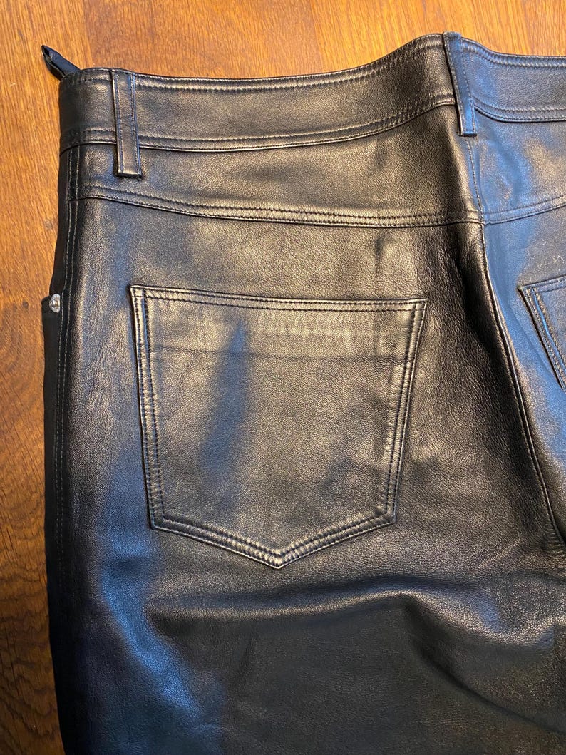 May include: Close-up of black leather trousers. The trousers have a classic five-pocket design with a back pocket visible. The leather appears smooth and has visible stitching details. The trousers have a belt loop and a button closure.
