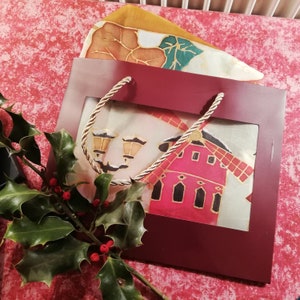 May include: A burgundy gift bag with a windmill design, featuring a red building with black accents and a white background. The bag has a gold and white rope handle and is decorated with a sprig of holly with red berries and a leaf-patterned scarf.