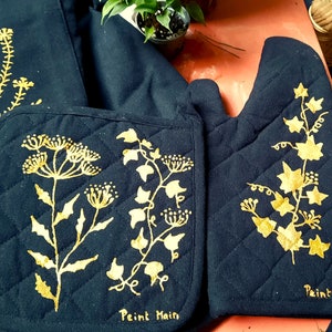 Könnte beinhalten: Ein Set aus marineblauen, gesteppten Küchentextilien, darunter ein Ofenhandschuh, ein Topflappen und ein Geschirrtuch, jeweils mit goldenen floralen Designs verziert. Die Worte "Peint Main" sind auf dem Topflappen und dem Ofenhandschuh aufgedruckt.