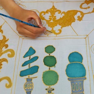 May include: A hand holding a paintbrush is adding gold paint to a white fabric. The fabric features a repeating pattern of teal and green topiary designs and gold floral accents. The artwork is in progress.