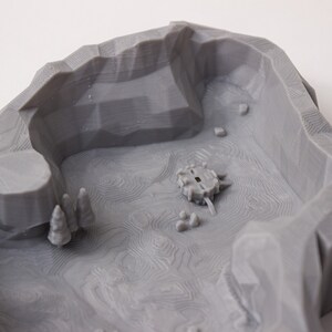 May include: A gray 3D printed model of a landscape with a small circular structure in the center. The landscape is textured with lines and has a small cave on the left side.