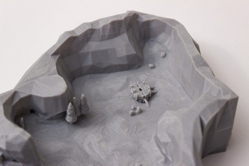 May include: A gray 3D printed model of a landscape with a small circular structure in the center. The landscape is textured with lines and has a small cave on the left side.