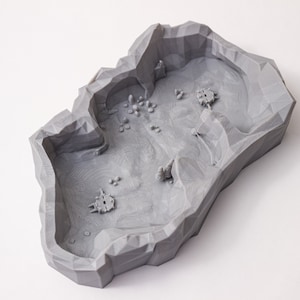 May include: A gray 3D printed terrain model with a rocky, cave-like design. The model features a central area with a rough, textured surface and several smaller, raised areas that resemble rocks or hills.