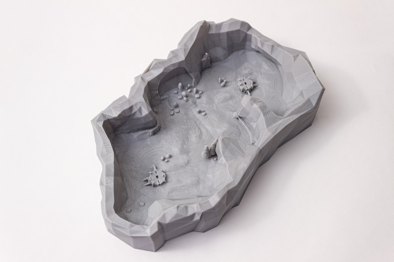 May include: A gray 3D printed terrain model with a rocky, cave-like design. The model features a central area with a rough, textured surface and several smaller, raised areas that resemble rocks or hills.