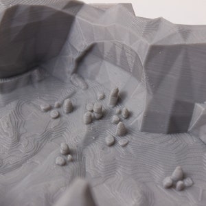 May include: A 3D printed model of a mountainous landscape with a gray finish. The model features a valley with several small, rounded peaks and a textured surface.
