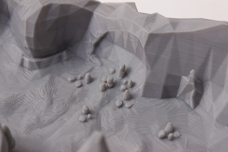May include: A 3D printed model of a mountainous landscape with a gray finish. The model features a valley with several small, rounded peaks and a textured surface.
