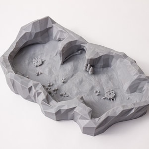 May include: A gray 3D printed terrain piece shaped like a rocky cave with a small opening. The cave is designed for tabletop gaming and has a textured surface.