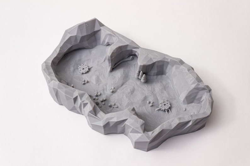 May include: A gray 3D printed terrain piece shaped like a rocky cave with a small opening. The cave is designed for tabletop gaming and has a textured surface.