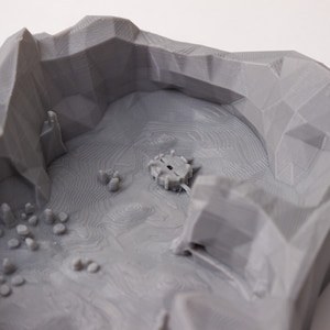 May include: A gray 3D printed model of a mountain range with a small structure in the center. The model is designed for tabletop gaming.