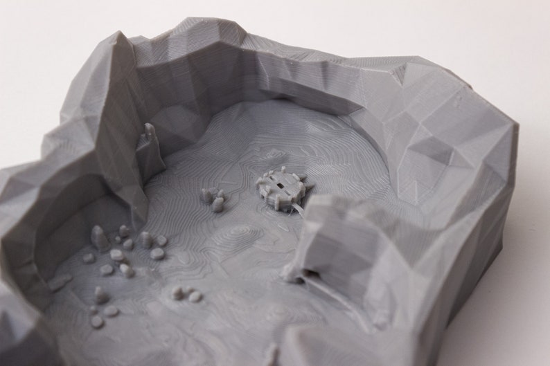 May include: A gray 3D printed model of a mountain range with a small structure in the center. The model is designed for tabletop gaming.