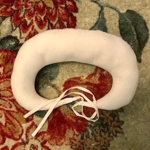 Könnte beinhalten: Ein weißes, oval geformtes Kissen mit einer zentralen Öffnung, gebunden mit weißen Bändern. Das Kissen ist aus weichem Stoff gefertigt und auf Komfort ausgelegt. Das Kissen liegt auf einem Teppich mit floralem Muster.