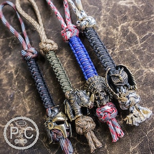 May include: Five colorful paracord keychains with different metal charms. The charms include a rhinoceros, a Native American headdress, an owl, and a Roman gladiator helmet.
