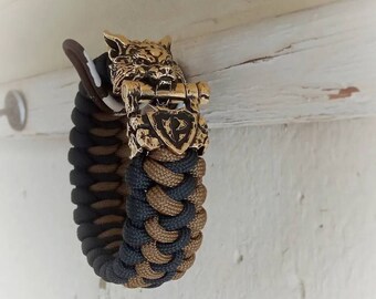 Puede incluir: Un brazalete de paracord negro y marrón con un cierre de cabeza de lobo. El cierre está hecho de metal y tiene un diseño detallado.