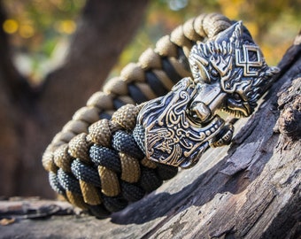 Puede incluir: Una pulsera trenzada de paracord en tonos beige, negro y gris, con un cierre detallado de cabeza de lobo plateado. La pulsera se exhibe sobre un trozo de madera texturizada, mostrando la artesanía.