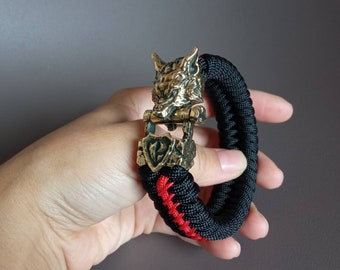 Puede incluir: Un brazalete de paracord negro y rojo con un cierre de cabeza de lobo de latón. La cabeza de lobo tiene un diseño detallado y una mandíbula articulada.