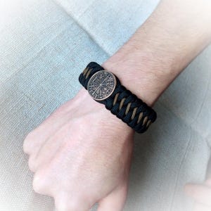 May include: A black and brown paracord bracelet with a round metal charm featuring a compass design and runes.