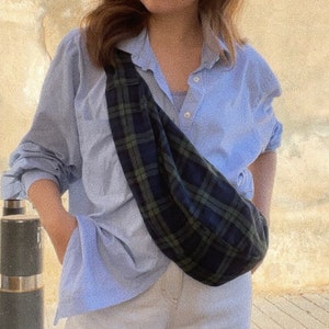 Puede incluir: Una persona lleva una camisa azul claro abotonada con una bolsa de bandolera de cuadros negros y verdes. La bolsa se lleva sobre un top blanco sin mangas y pantalones blancos.