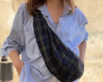 May include: A person wearing a light blue button-down shirt with a black and green plaid crossbody bag. The bag is worn over a white tank top and white pants.