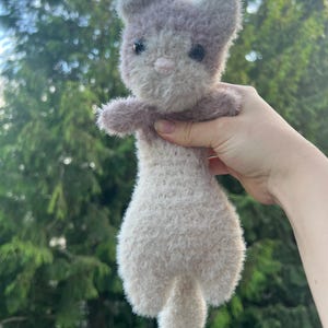 May include: A handmade plush cat toy in shades of grey and white. The cat has a fluffy texture, small black eyes, and a pink nose. The toy is held up against a backdrop of green foliage.